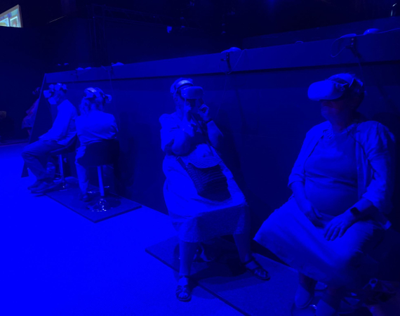 Three older adults test VR headsets in a darkened, blue room.