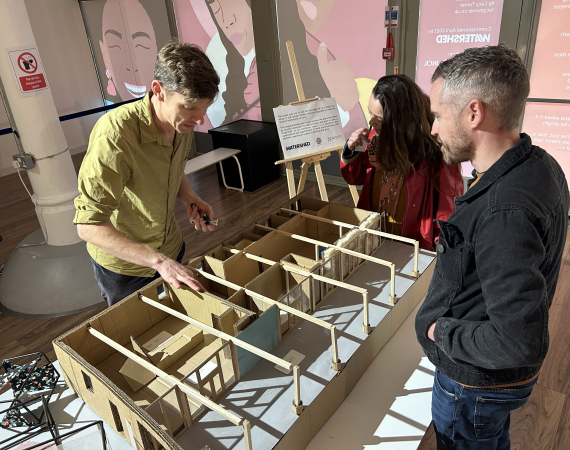 Three people gather around a wood and cardboard model of Undershed, the Watershed's new immersive gallery space.