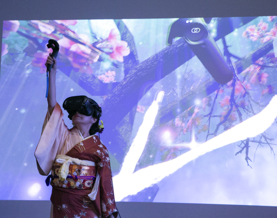 A woman dressed in traditional Japanese clothing, in a VR headset, plays with a VR performance piece, projected onto a screen behind her. 