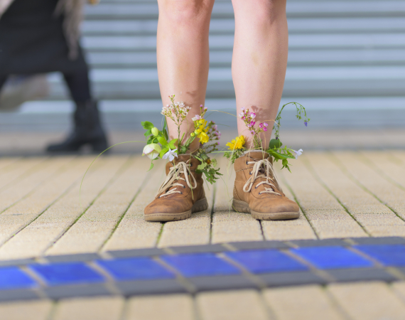 A photo of a pair of legs from the knee’s down, with the feet adorned in brown boots brimming with flowers. In the background you can see the silver shutter of a shop and the legs of another person walking past.
