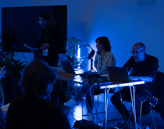 An image of a tranquil space, with an audience to the left, and three sound artists to the right, sat at a table, sharing a work of sound art. The room is lit in a peaceful, dark blue.
