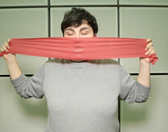 An image of a person, eyes closed, wearing a grey jumper, in front of a tiled background. They are stretching a piece of red rubber over their open mouth, creating an impression in the rubber.