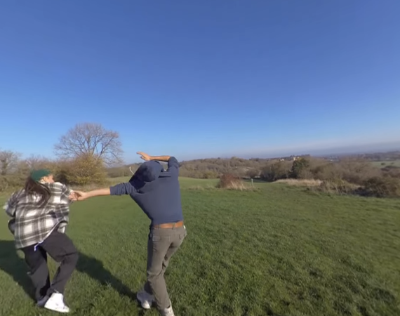 A screen capture from a 360 image of two people playfully moving around in a lush green space with a clear blue sky.