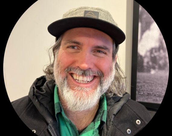 An image of Kyle Keane, a white, bearded man, smiling at the camera. He wears a flat peak cap, black jacket, and green flannel shirt.