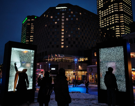 An image of a city at night, with two light fixtures in the foreground, on which human figures are silhoutted, interacting with the fixtures as part of a sensory exhibition.