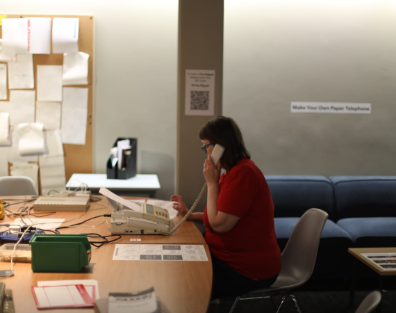An image of Frances Bossom in her 90's style office installation at the arnolfini. She sits at a table, with an old corded phone to her left ear - on the table is a fax machine, and various pieces of paperwork. The wall in the background has a noticeboard covered in paper notices.
