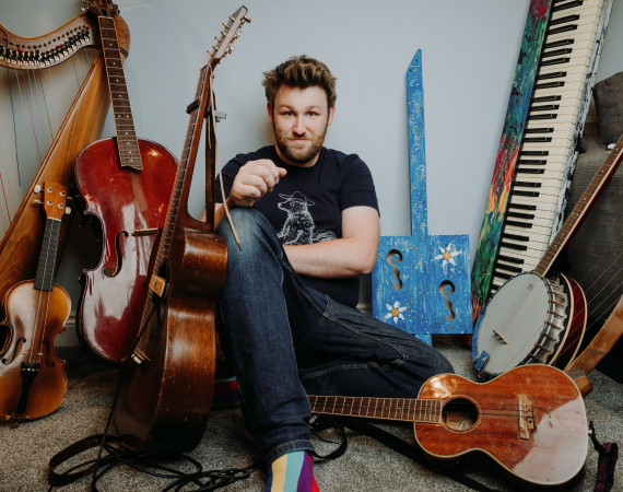 An image of the artist and creative, Greg Ryan, a white man in his mid to late 30's, sat amongst a vast array of predominantly acoustic instruments. He is looking directly into the camera, and wears blue jeans and a navy blue t shirt.