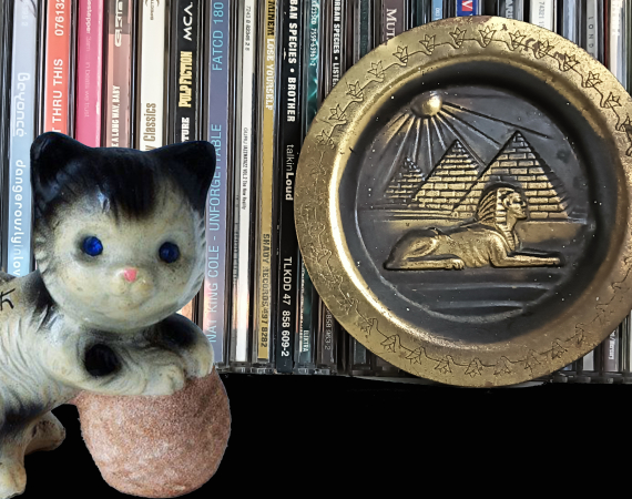 An image of overlaid ornaments in front of CDs on a shelf - one ornament is a china cat playing with a ball of wool, and the other is a brass bowl depicting a sphinx and the pyramids of egypt.