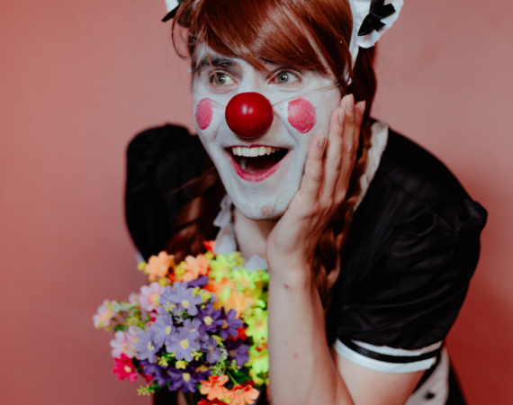 An image of Alistair Aitcheson in an androgynous clown/maid costume; he is cupping his head with his left hand with a bashful, cheery expression.