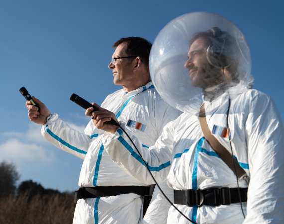 An image of two men, side by side, dressed in white uniforms that resemble astronaut uniforms. The man on the right, musician Mr. Kit, wears a fishbowl-like helmet on his head. Both men are extending their right arms outward, and are holding microphones with which to record their environment.