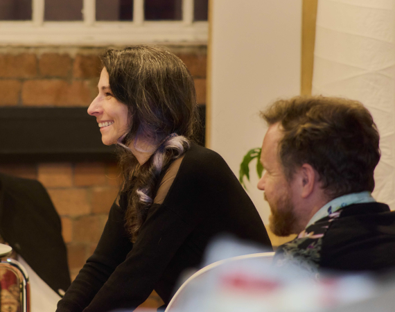 Two white people are smiling over to the left of the screen, they're both sat down.