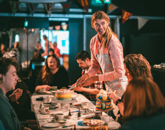An image of Tom Marshman's tea party - Tom is standing, looking off camera to the right, about to cut a rainbow coloured cake sat on a white table, surrounded by happy participants.
