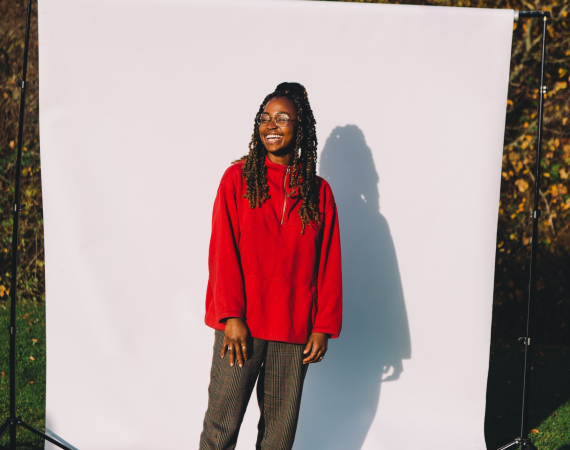 An image of a Emmanuella Blake-Morsi, against a white backdrop situated in an outdoor green space. Emmanuella is a black woman, wearing glasses, a red sweater and dark green trousers, looking off camera to the left, with a smile on her face. 