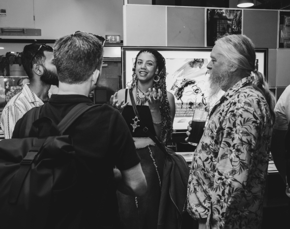 A group of five people in a lively discussion in an indoor setting. A woman in the center gestures expressively, flanked by conversational companions. The mood is engaged and friendly.