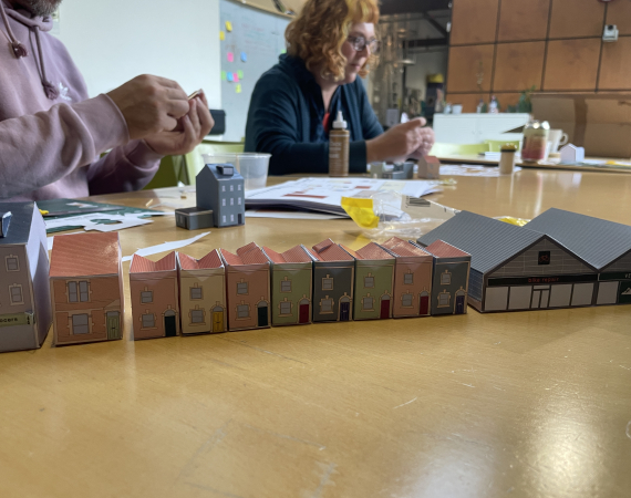 Two people sit at a table assembling small, colorful paper model houses. The atmosphere is focused and creative, with a variety of materials spread out.