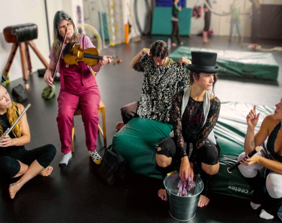 A group of circus performers, all white women, sit in a semi circle in a rehearsal space with a black floor. Some sit on the floor, while others sit on green cushions or chairs. They are in discussion, while two performers on the left play instruments - one, on the far left, plays the flute, while the other to her right plays a fiddle. 