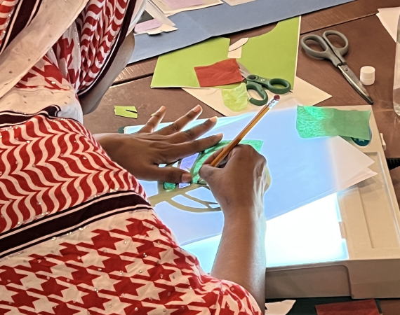 A person in a red and white patterned headscarf creates art with paper and scissors on a lightbox at a craft table, conveying focus and creativity.
