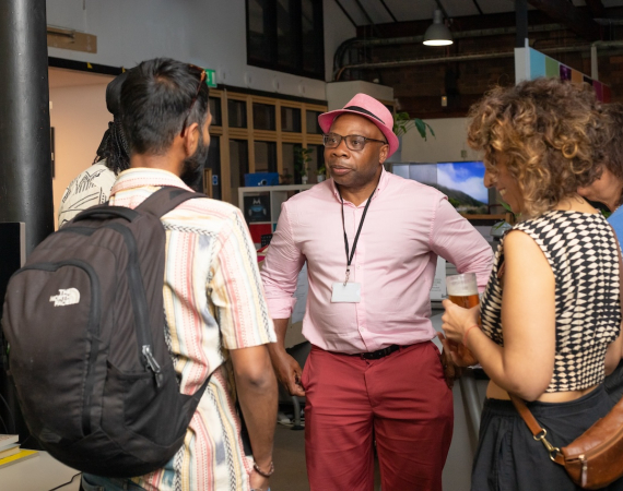 A group of four people stand in an office space, engaged in conversation. One person in a pink hat and matching trousers appears to be speaking. The atmosphere is casual and sociable.