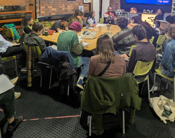 A diverse group of people sit around a rectangular table in a modern meeting room with exposed brick walls. A presentation titled "Key Objectives & Messages" is projected on a screen. The room is well-lit with string lights, creating a collaborative and focused atmosphere.