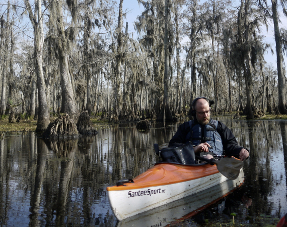 A man in a life jacket and headphones kayaks through a calm, mirror-like swamp, surrounded by tall cypress trees draped in Spanish moss, conveying tranquility.