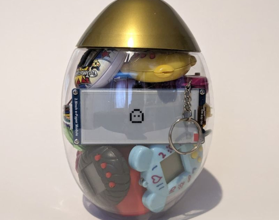 A transparent egg-shaped container with a gold top holding small toys, including a digital pet keychain and a smiling emoji screen, against a plain background.