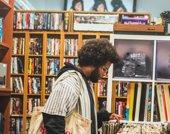 An image of the musician Lawi Anywar a Black man with an afro is browsing vinyl records in a vintage market. He is surrounded by various types of physical media, and carries a white tote bag full of records. 