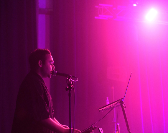 An image of Nik Rawlings singing into a microphone. There is a laptop in front of them, and the room they are performing in is illuminated entirely in a pinkish, purple hue. Nik wears all black and is prominent to the left of the image. 