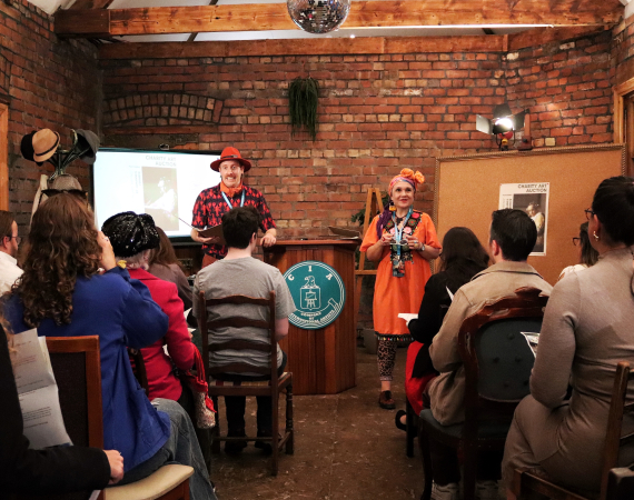 An image depicting Paul (on the left in red) and Gill (on the right in orange) delivering a theatre experience in a red brick room with a smartly dressed audience. They are flanked by a cork board on the right and a small projection set upon the left. 