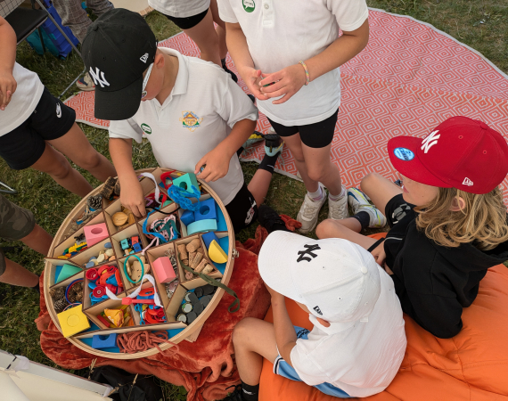 An image of young people gathered around a container of craft materials as they deliberate how to use them. Two of them are sat down on an orange covered seat on the right, and four more stand in the center of the image, interfacing with the container of craft materials. 