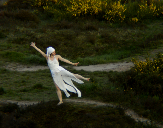 An image of Anastasia Dahl, dressed entirely in white, performing a piece of movement in an outdoor earthy green space - she is surrounded by yellow flowering gorse bushes to the top and right of the image. She is stood on one leg, with her left arm and leg pointing outward, and her right arm pointing upward. Her head faces the right of the image. 