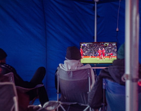 An image of men watching TV in a refugee camp. The TV shows a football match. 
