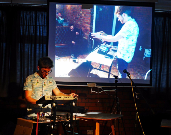 Ant Nasce a white man with glasses on stands at a Korg synthesizer, performing music under dim lighting. A large screen behind him displays a projected image of another person adjusting equipment, conveying a creative and focused atmosphere.