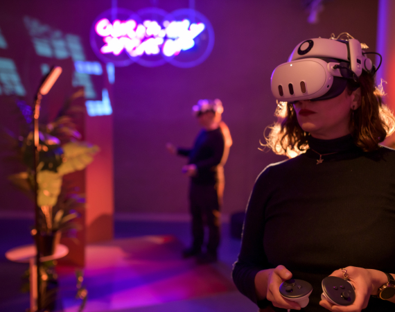 Person standing with a VR headset against a brightly coloured environment with another person in a headset in the background