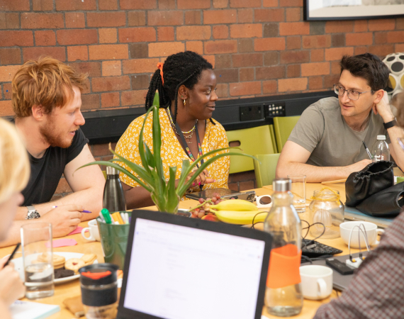 Five people engaged in a friendly discussion around a table with laptops and drinks. A plant adds greenery. The setting is casual and collaborative.