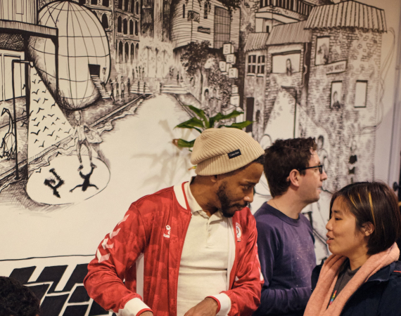 A man plays video games at a round table, while a person in a red jacket and beanie talks to a smiling woman. The background features a detailed mural.