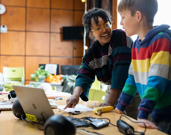 Smiling woman and boy collaborate at a table with a laptop and craft supplies in a bright classroom, creating a warm, engaging scene.