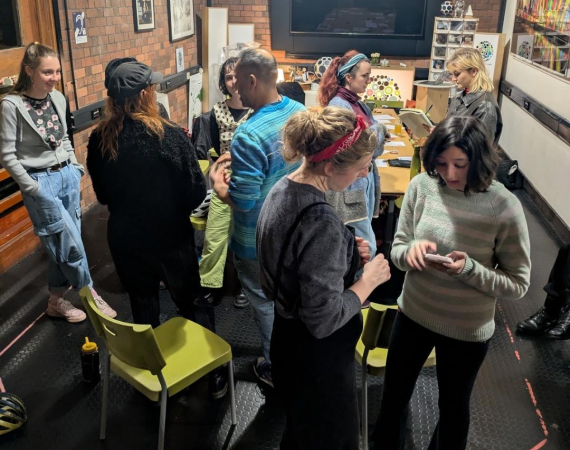Adults gather in a cozy brick-walled workshop with string lights, chatting around tables and chairs; a screen and colorful artwork create a creative mood.