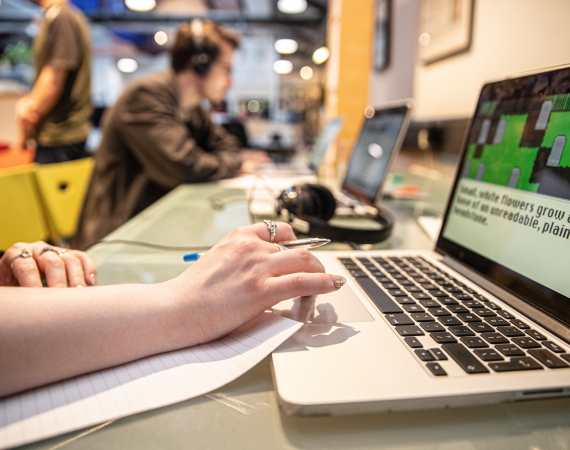 An office work place scene. In the foreground a hand rests on an open laptop holding a pen and paper. In the background out of focus another person sits at a desk wearing headphones 