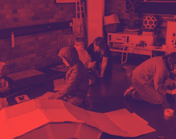 In a red-tinted workshop, several people crouch around large sheets on the floor, focused on a project beside equipment and circular artwork.