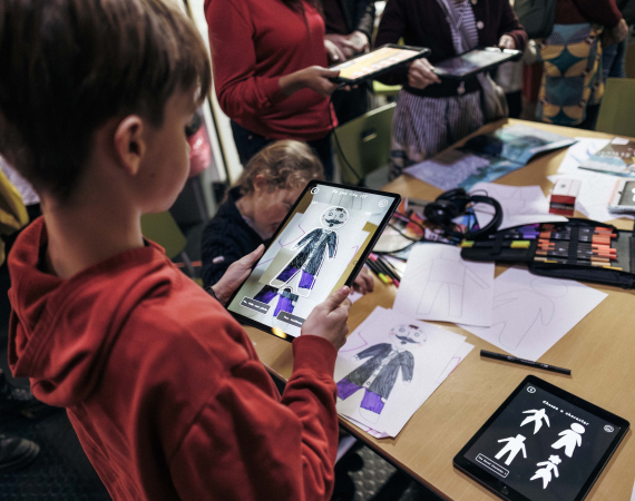 A child in a red hoodie uses a tablet to design clothing at a busy fashion workshop, surrounded by sketches, fabric swatches, and other participants.