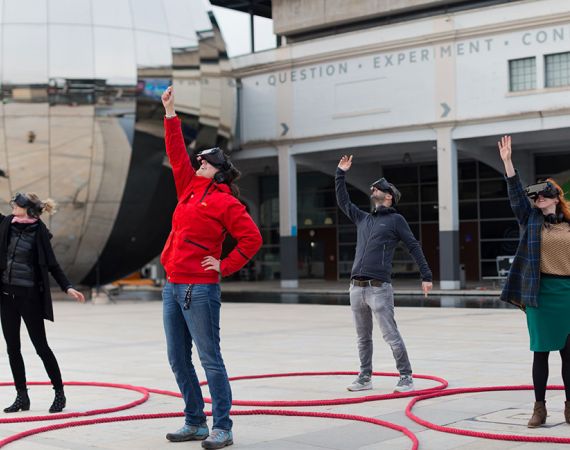 Four people in VR headsets with synchronised arms in the air experiencing From The Light Of The Fire, Our Dancing Shadows.