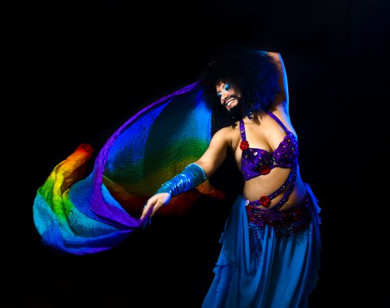 Image Shrouk El-Attar belly dancing in drag, with a rainbow coloured piece of cloth mid dance.