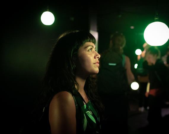 An image of a person with long black hair and a short fringe looking into the distance in a dark room, they are lit up in green neon light, and there are bright light bulbs scattered around in the background 