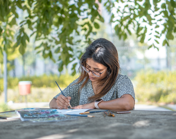 A woman in glasses paints on a table outdoors, under a tree with leaves in the background.