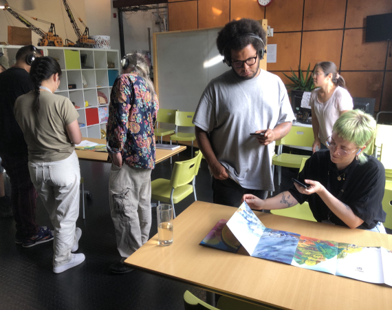 Image of seven people in the Pervasive Media Studio events space. A group of two are facing the camera and intently looking down at a book. Another group of four people, with backs to the camera, are also looking down at another copy of the same book. Another person standing on their own in the background looks into the distance.