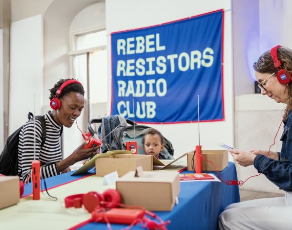 A young Black women sits smiling with red headphones on and a banner above her reads Rebel Resistors Radio Club. 