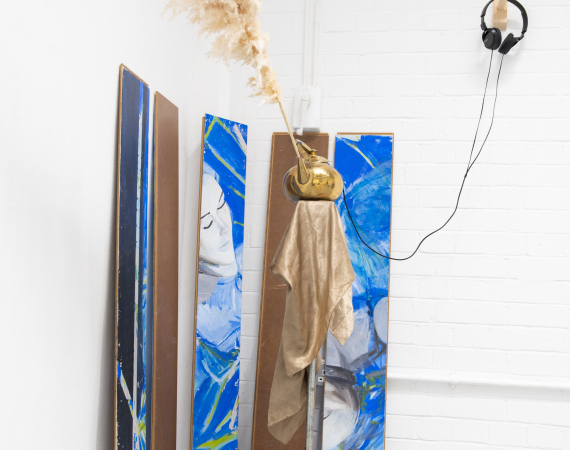 Art installation with pampas grass in a gold vase on a stand, draped with metallic fabric. Three abstract panels in blue and brown lean against a white wall. Headphones hang nearby.