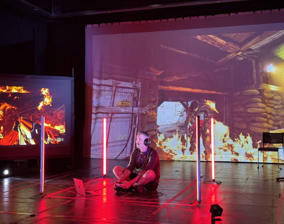 A person sits cross-legged on the floor, wearing headphones, surrounded by vertical red lights. Intense video game scenes with fire project in the background.