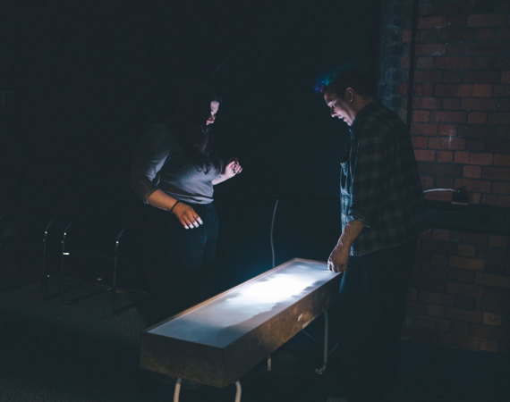 Two people stand in a dimly lit room, focused on a glowing rectangular table. The scene conveys curiosity and concentration against a brick wall backdrop.