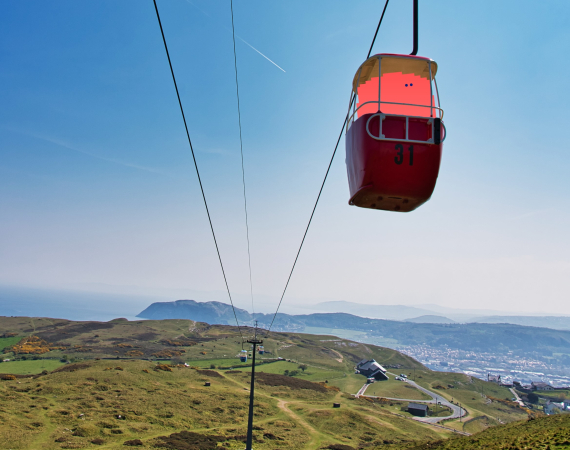 A coral coloured barrel like character sits in a cable car with a beautiful landscape behind it in Llandudno.